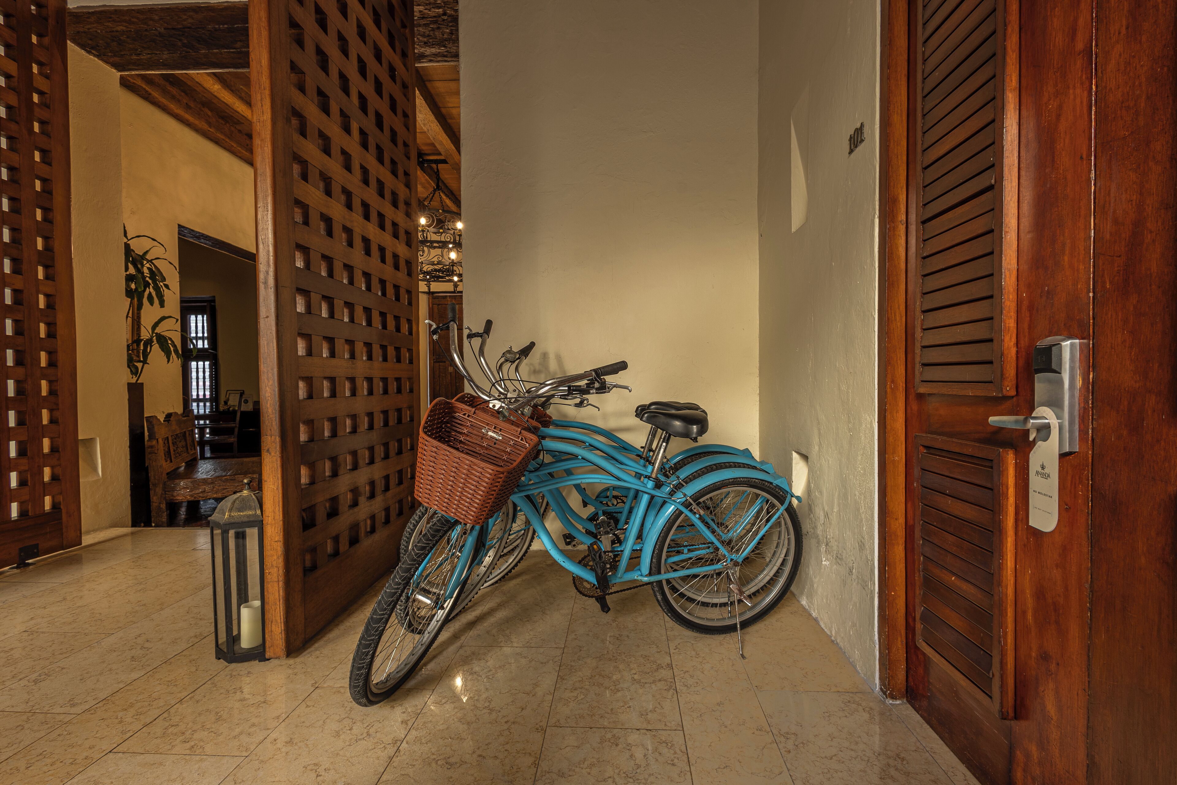 A blue bicycle with a basket and a lantern, leaning against a wooden door in a hallway with tiled flooring.