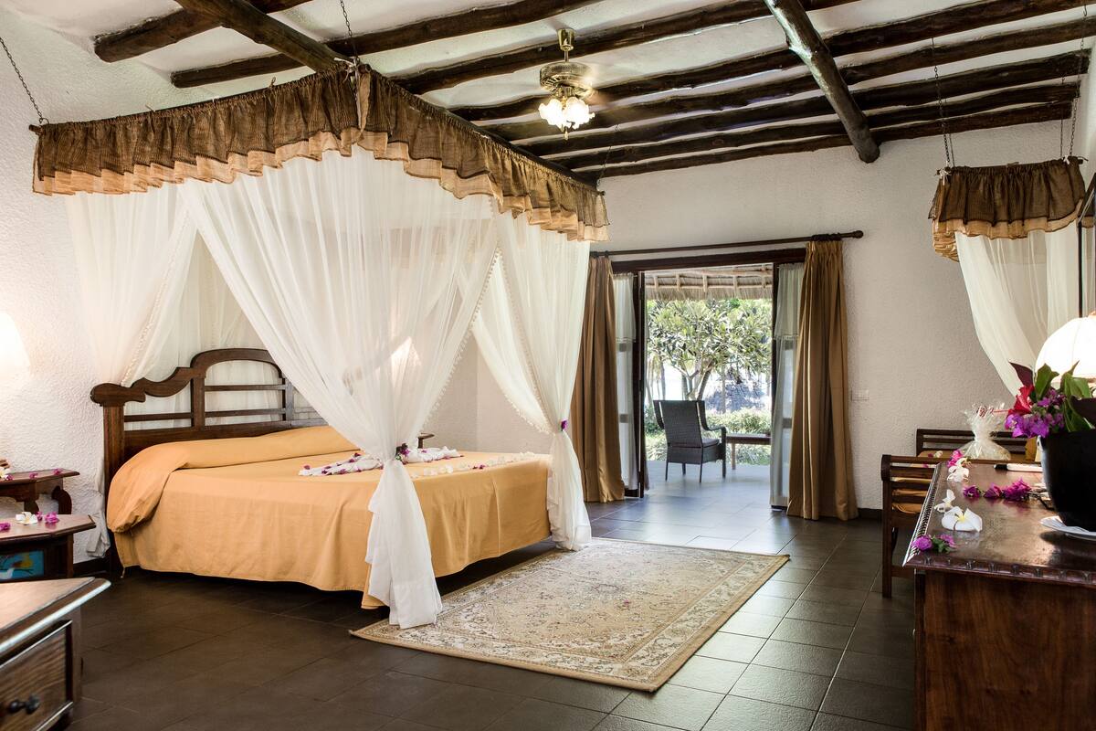 A bedroom with a bed, a wooden headboard, a nightstand, a desk, and a chair. There is a window with curtains, a door leading to an outdoor area, and a ceiling fan.