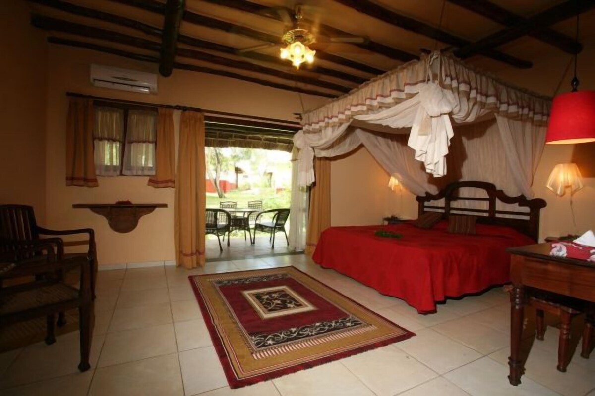 A bedroom with a canopy bed, a red bedspread, a wooden headboard, a red lamp, a wooden nightstand, a wooden chair, a small table, a decorative rug, a window with curtains, and an air conditioning unit.