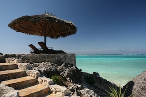 A stone staircase leading to a beach area with a thatched umbrella and lounge chairs.