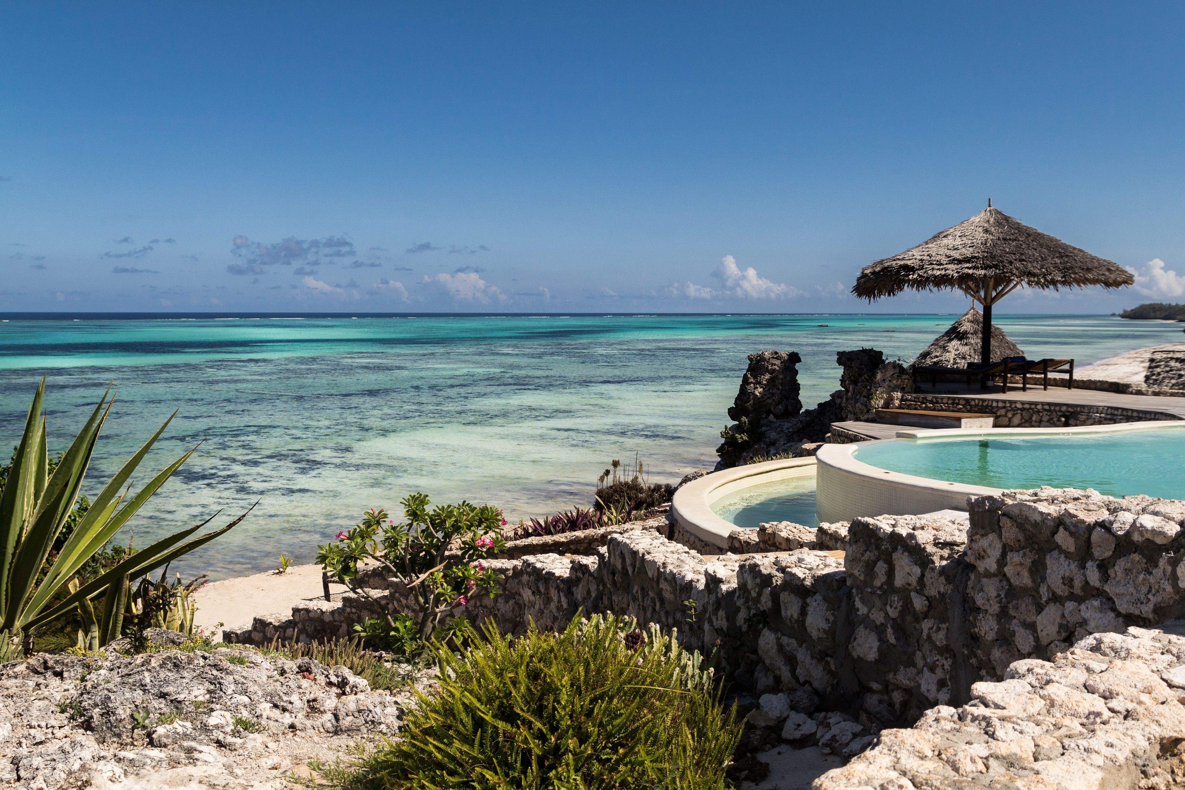 A pool area with a thatched umbrella, surrounded by stone walls and tropical plants.