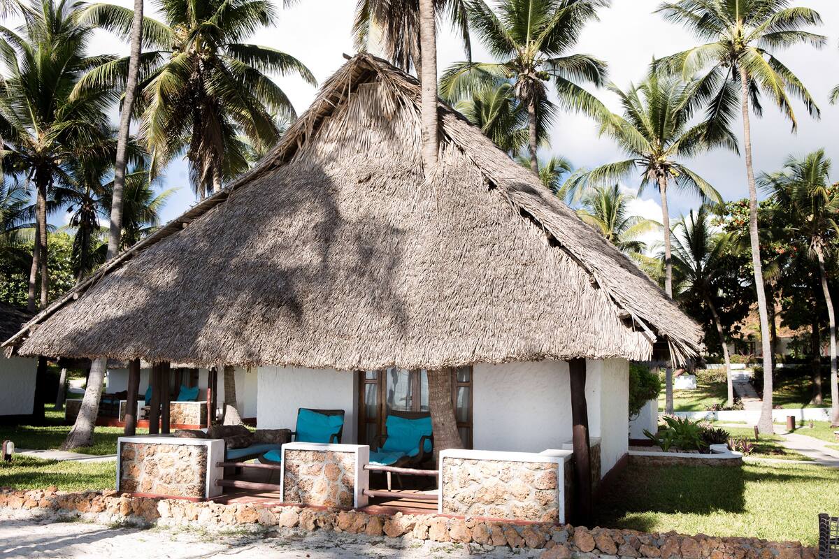 A thatched-roof hut with a stone wall, blue cushioned seating, and a small garden area.