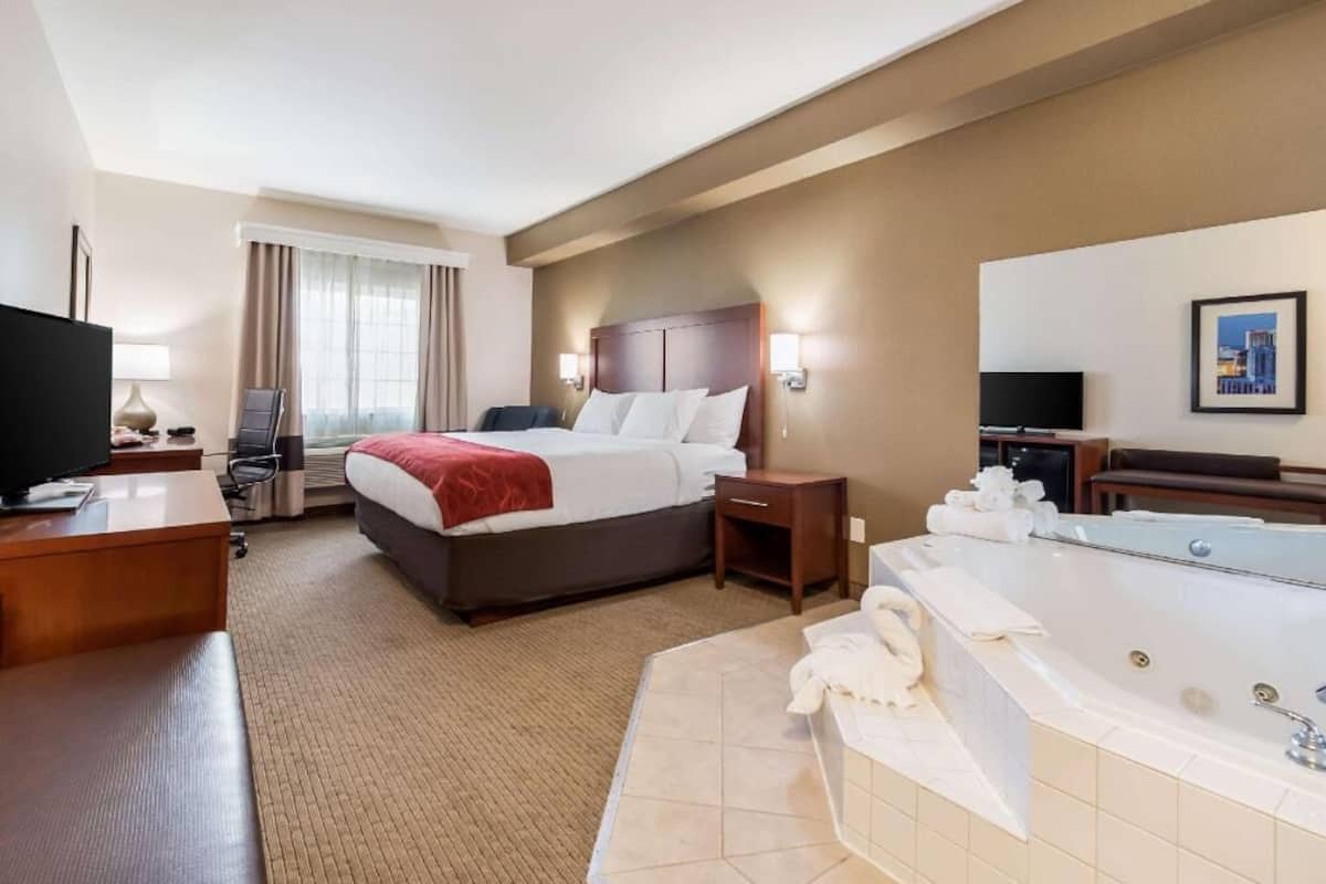 Habitación de hotel con cama, escritorio, televisor, baño con bañera y un ventanal con cortinas.