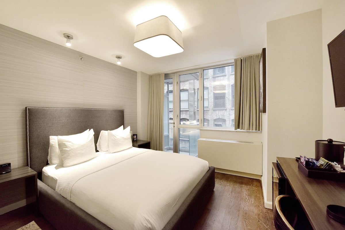 Un cuarto de hotel con una cama grande, una mesita de noche, un escritorio, una ventana con cortinas y vistas a la ciudad.