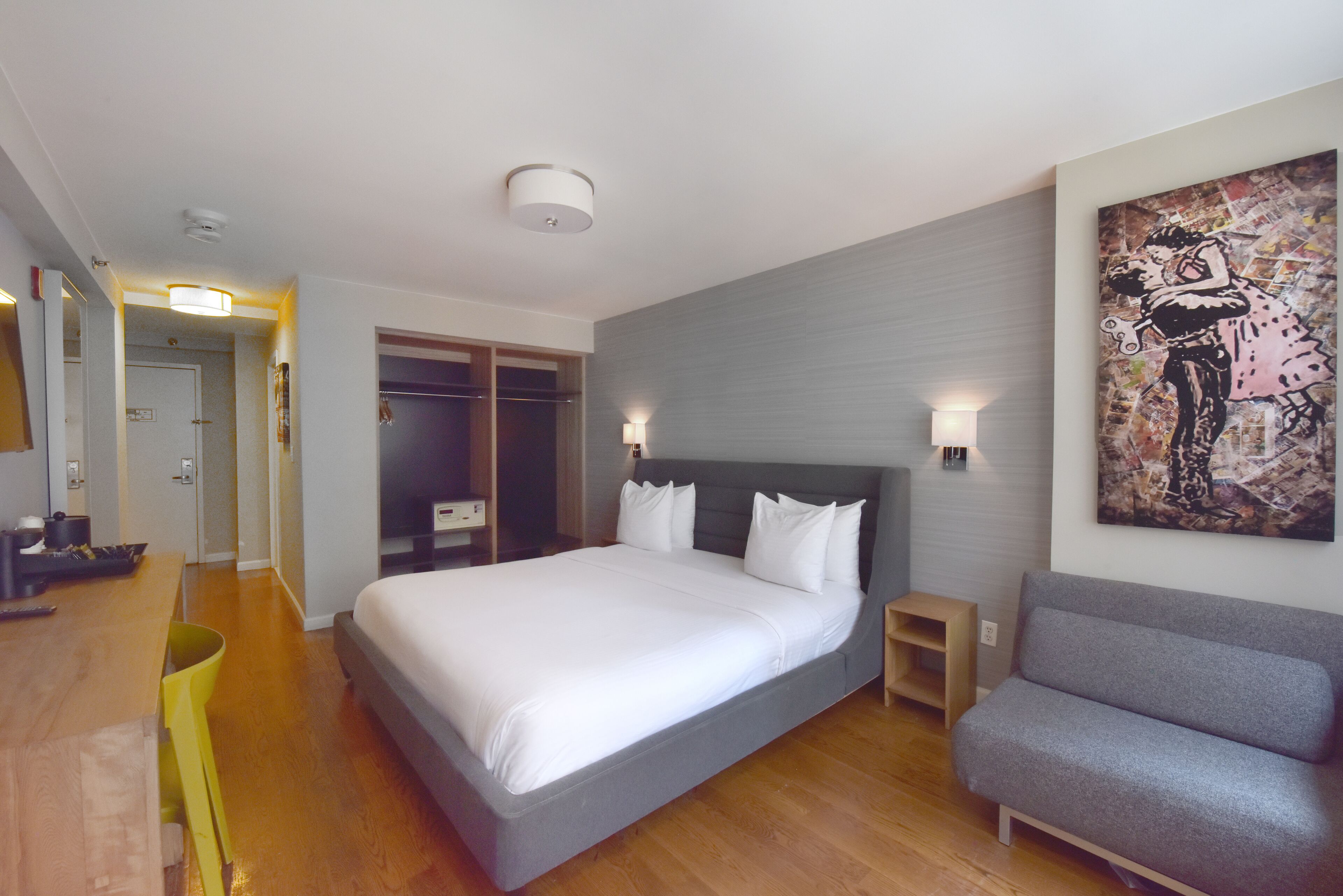 Habitación de hotel moderna con una cama grande, un sofá gris, un cuadro en la pared y un pasillo que conduce a un baño.