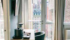 A modern hotel room with a wooden desk, a black chair, a bed, and a large window offering a city view. - Hotel 32 32 (New York)