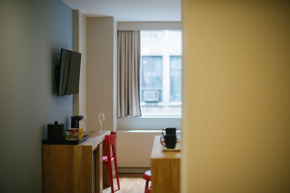 Una habitación pequeña y moderna con un escritorio de madera, una silla roja, un televisor montado en la pared y una ventana con cortinas.
