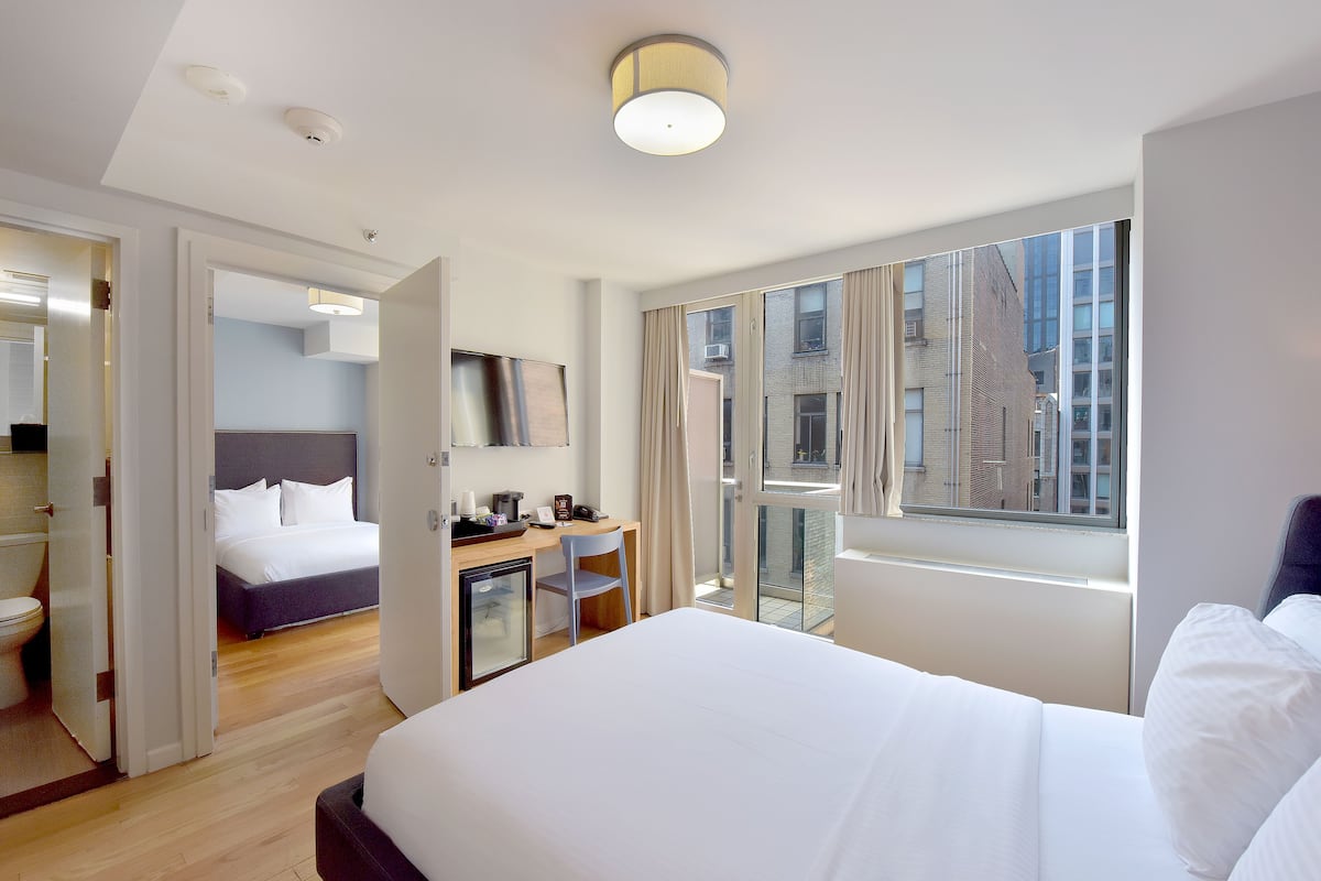 Una habitación de hotel moderna con una cama grande, un escritorio con silla, un refrigerador pequeño y un balcón con vista a la ciudad.