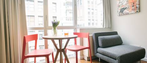 A room with a sofa, two chairs, a small table with a vase and a glass, and a window with curtains.