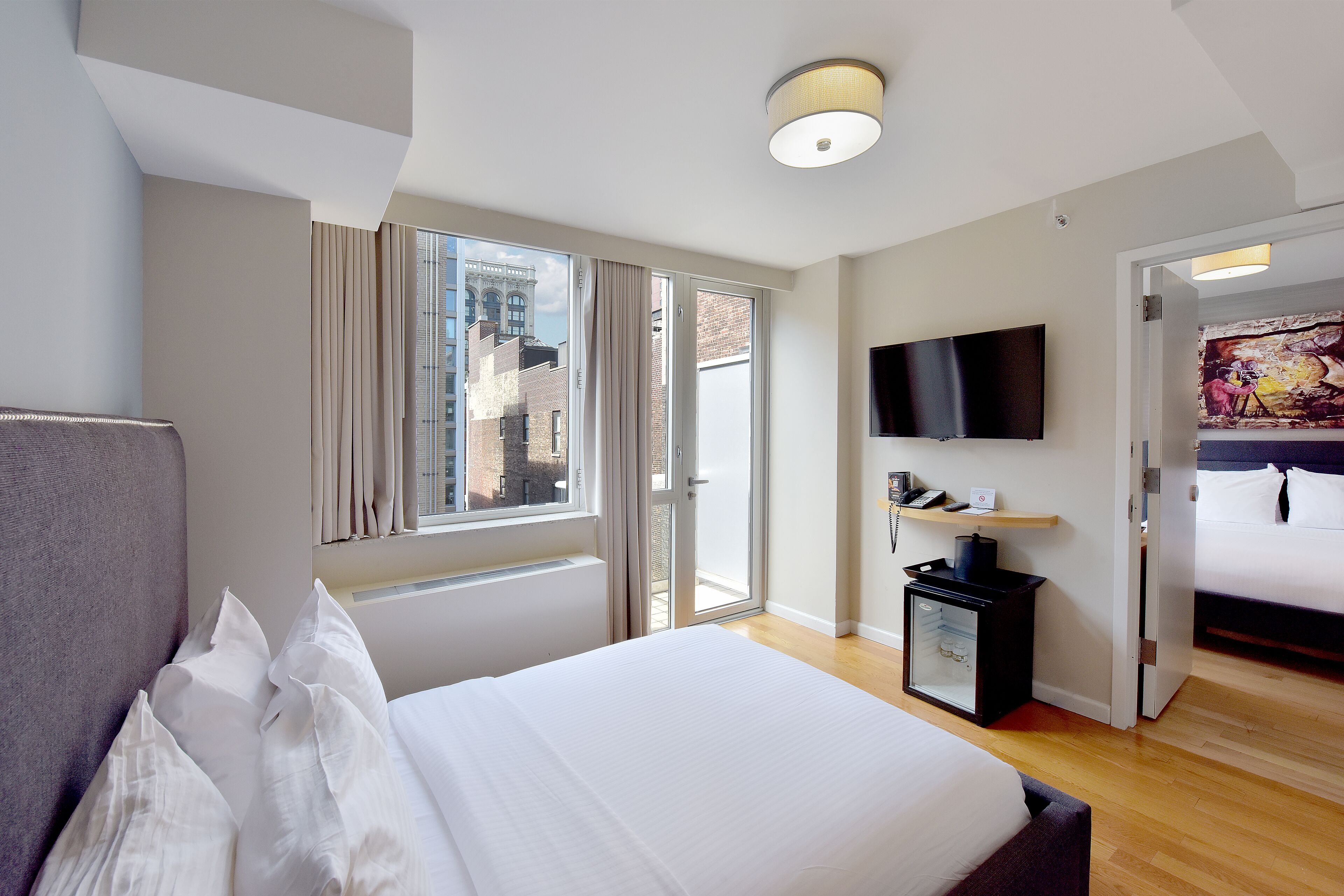 Habitación de hotel moderna con una cama grande, un televisor de pantalla plana en la pared, un escritorio pequeño con una lámpara y una puerta corrediza de vidrio que da a un balcón.