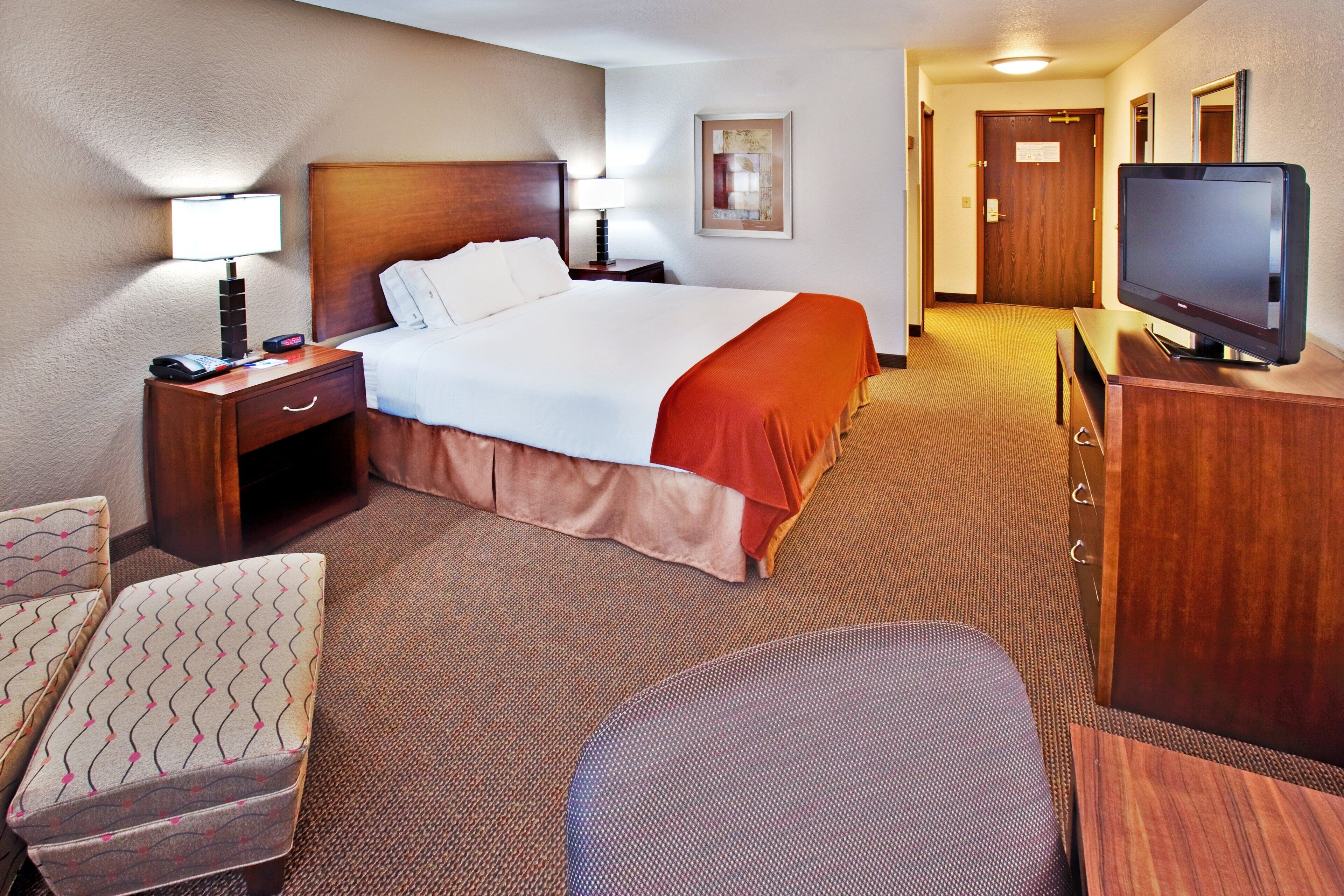 A hotel room with a bed, a television, a desk with a lamp, a chair, and a carpeted floor.