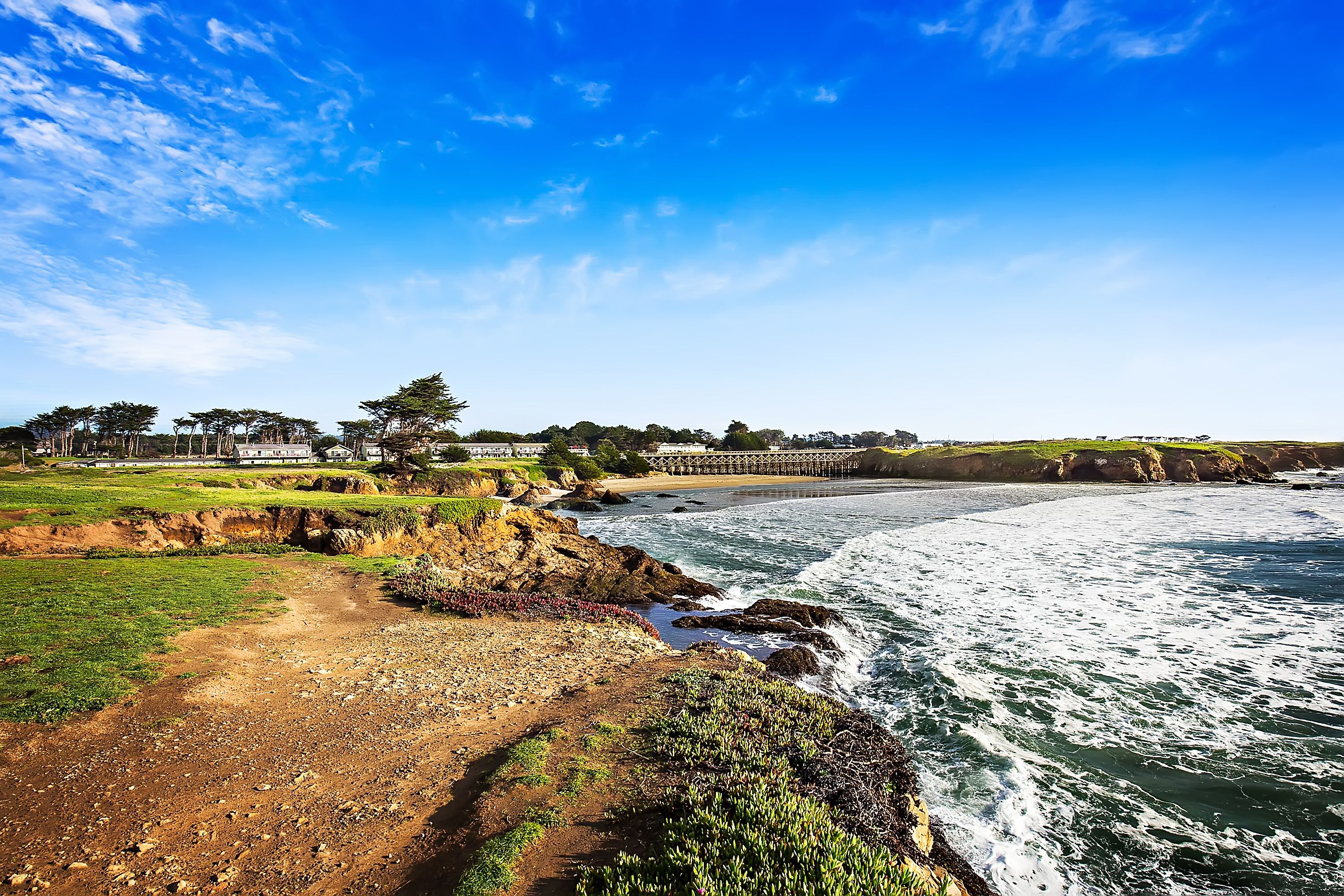 A coastal landscape with a rocky shoreline, a bridge, and a row of houses.
