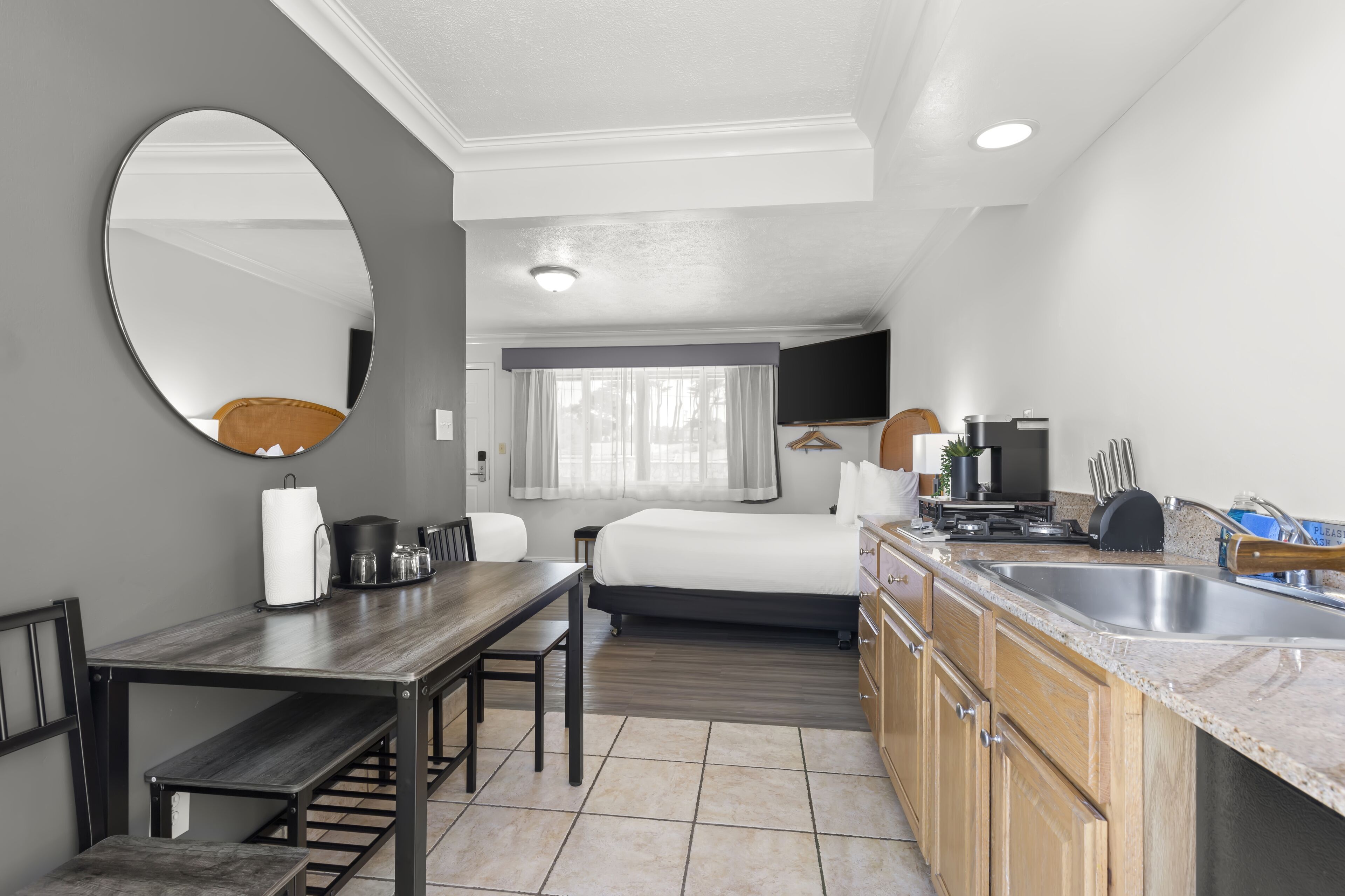 A compact hotel room with a bed, a dining table, a kitchen area with a sink, and a large mirror.