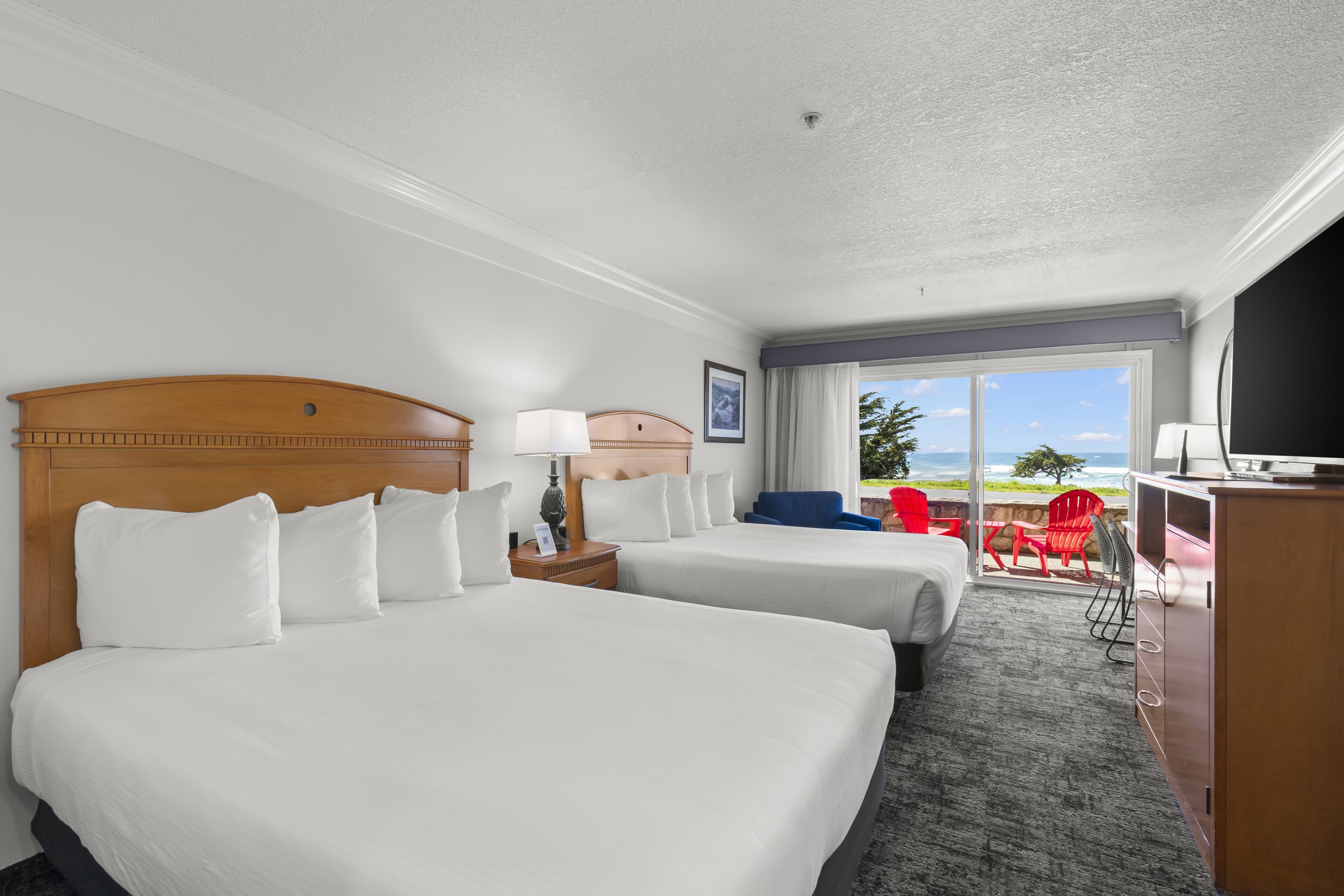A hotel room with two beds, a desk, a TV, and a balcony with red chairs.