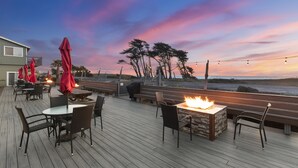 A wooden deck with outdoor seating, a fire feature, and a view of the ocean at sunset.