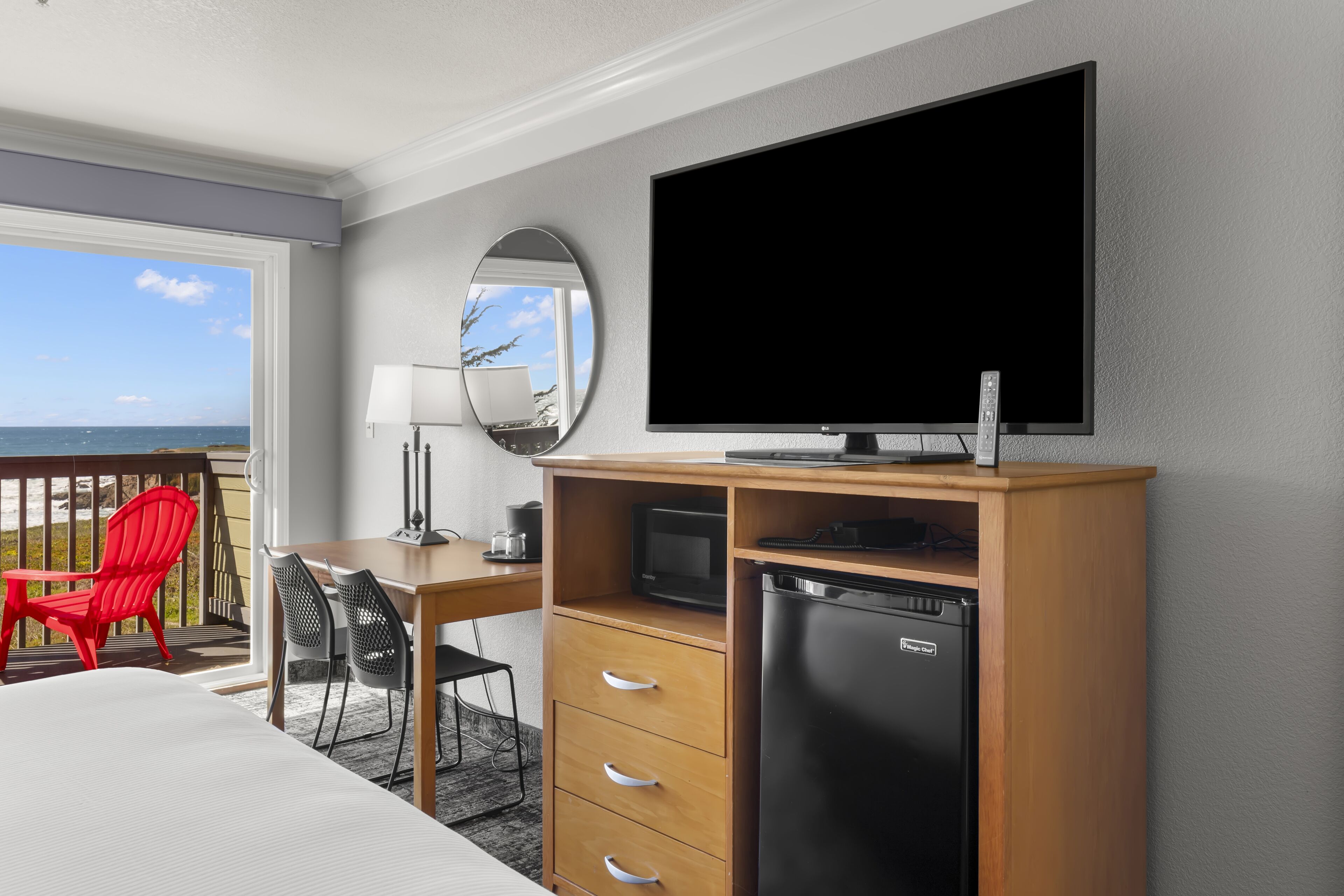 A hotel room with a bed, a desk with a chair, a television, a refrigerator, a microwave, and a view of the ocean.