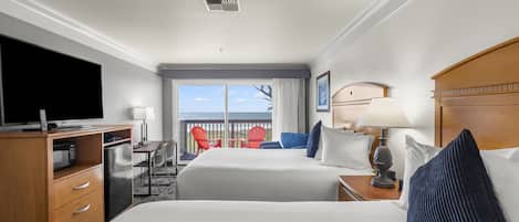A hotel room with a large bed, a desk with a computer, a TV, and a balcony with red chairs.