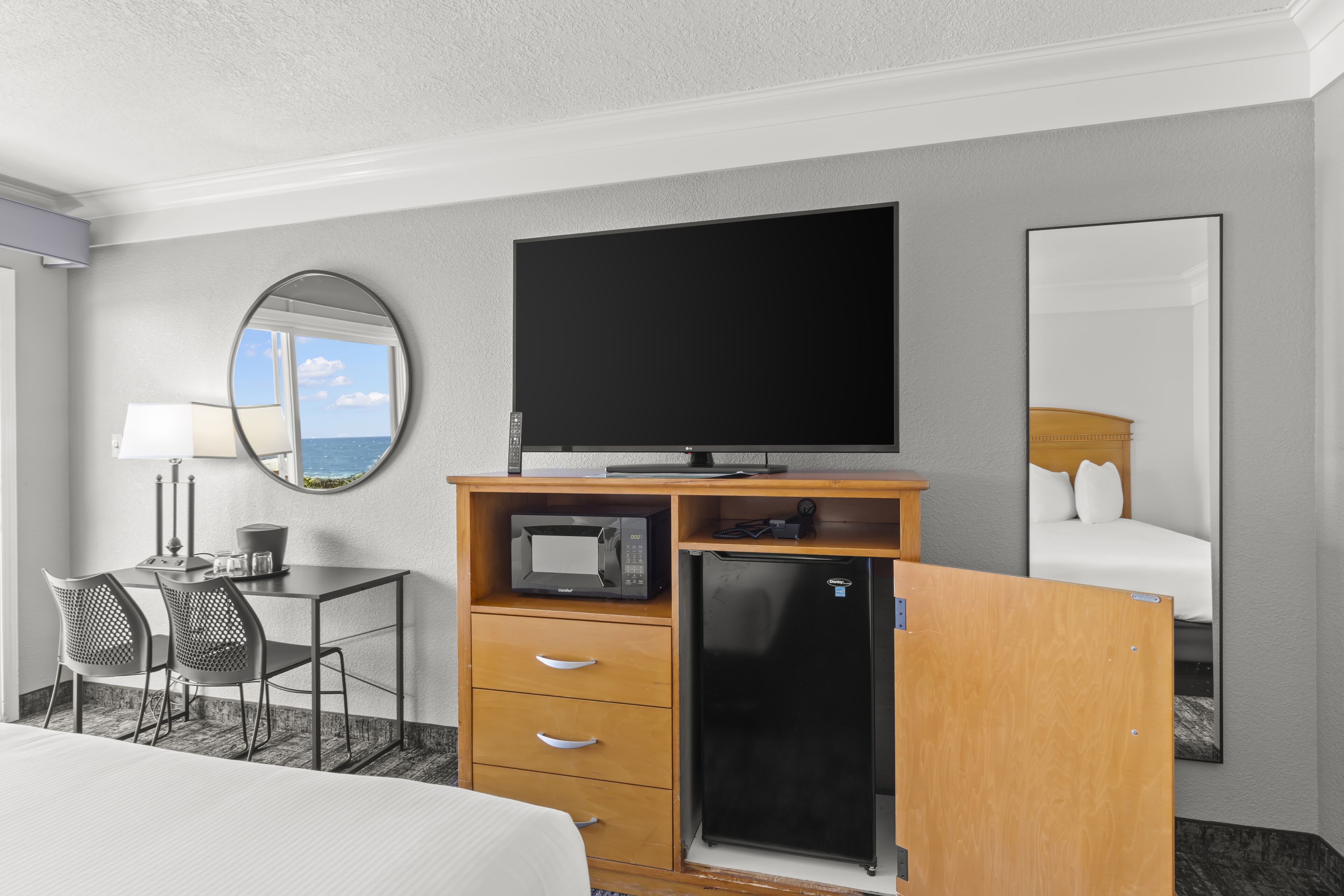 A hotel room with a bed, a desk with a chair, a television, a refrigerator, and a mirror.