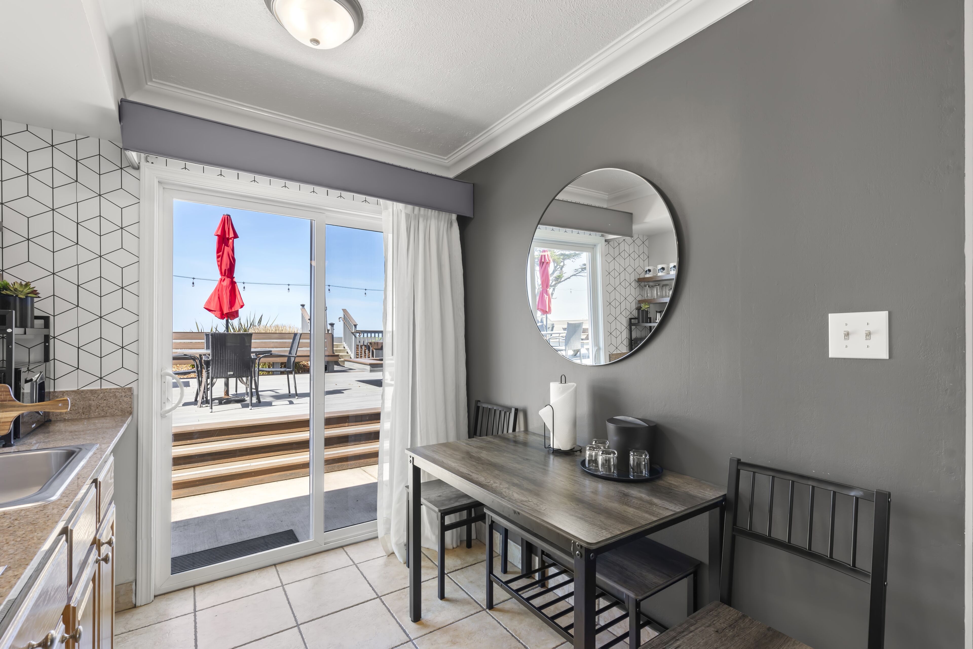 A modern kitchen with a dining area, a large window with curtains, and a round mirror on the wall.