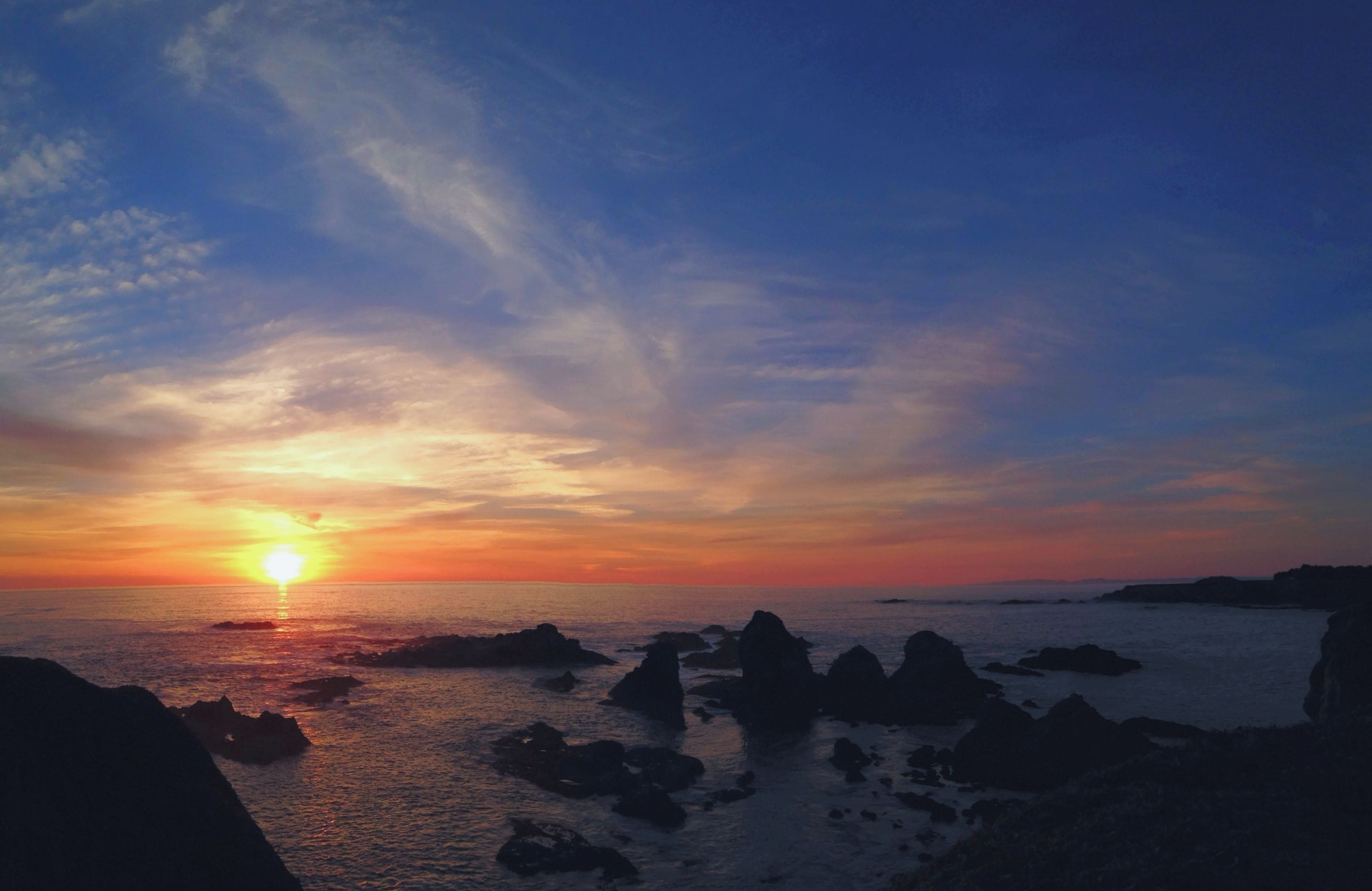 A sunset over a rocky coastline with the sun visible near the horizon.