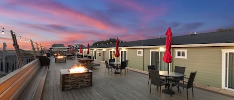 Terraza de madera con mobiliario exterior, una chimenea y sombrillas rojas.