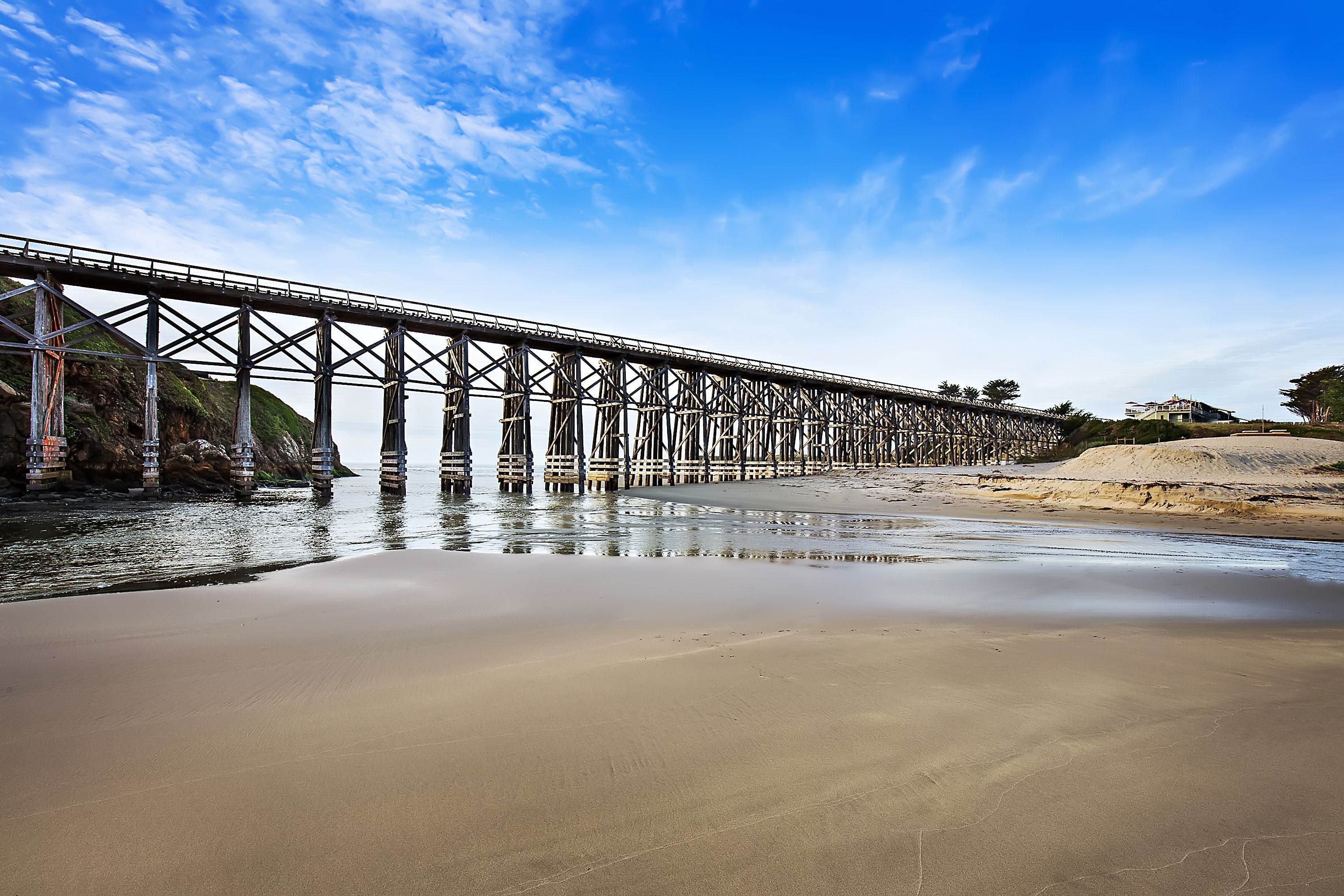 Un long pont métallique enjambant une étendue d’eau calme, avec une plage de sable au premier plan.