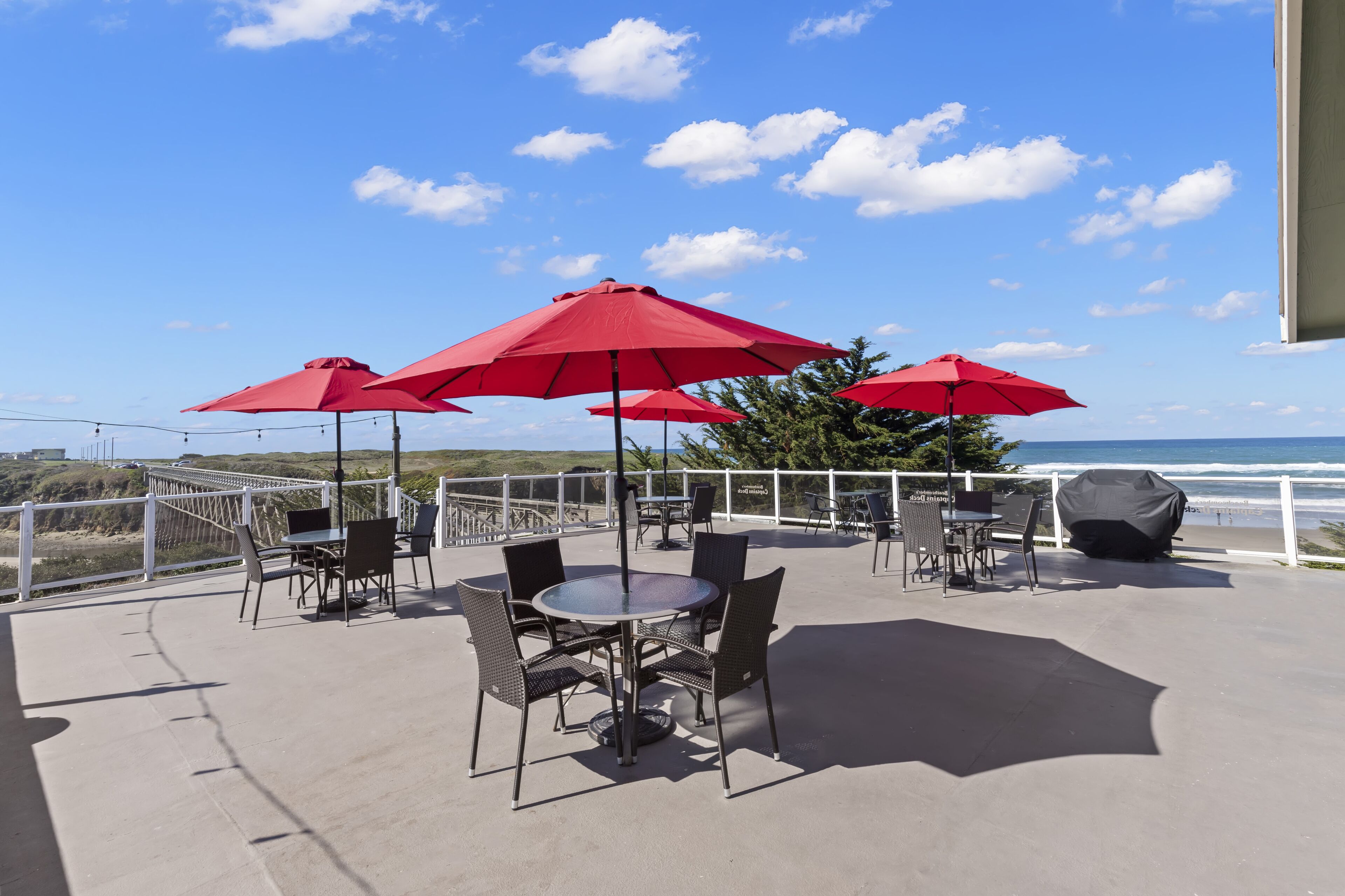 An outdoor patio with round tables and chairs, red umbrellas, and a view of the ocean.