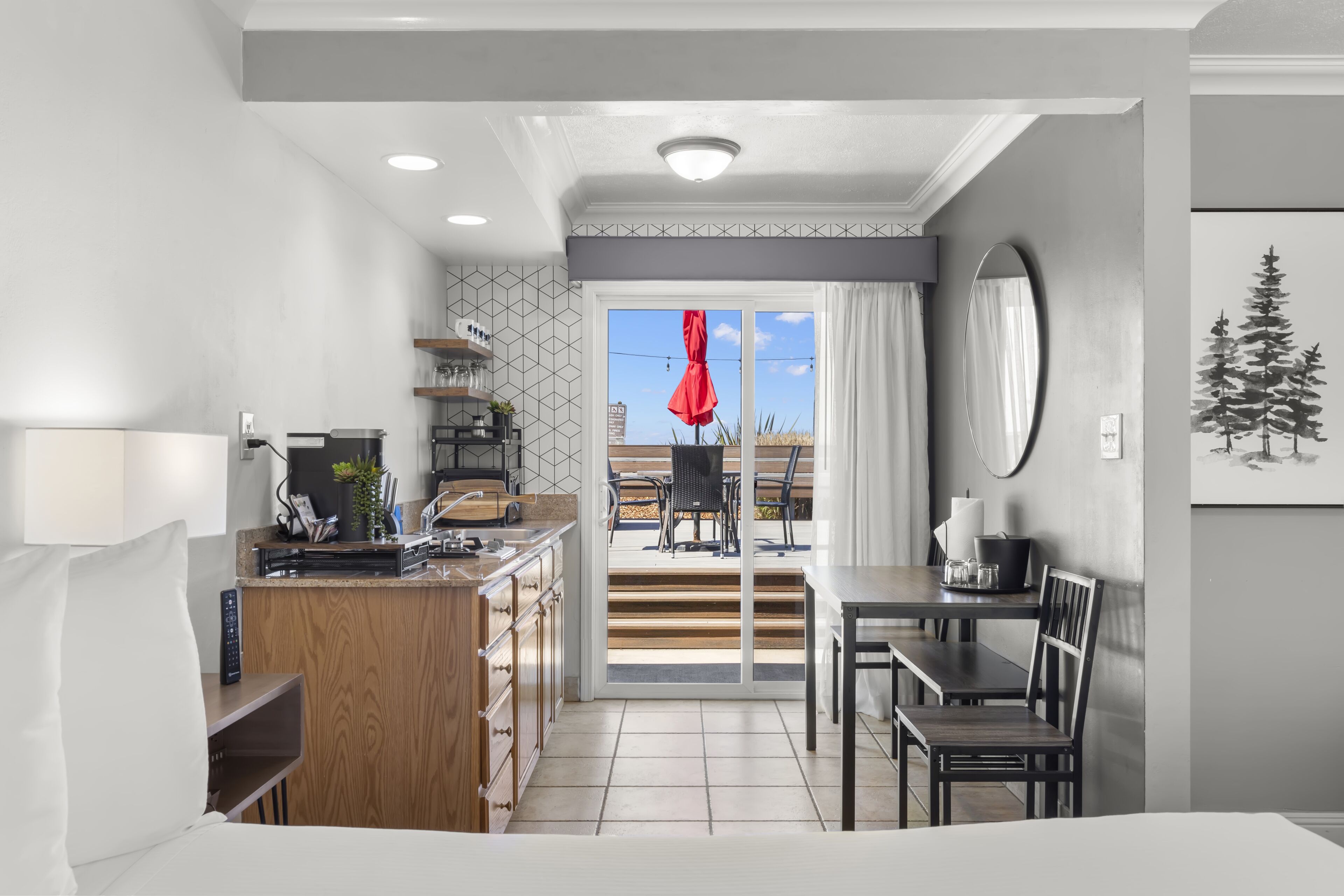 A modern hotel room with a kitchenette, a dining area, and a sliding glass door leading to an outdoor patio.