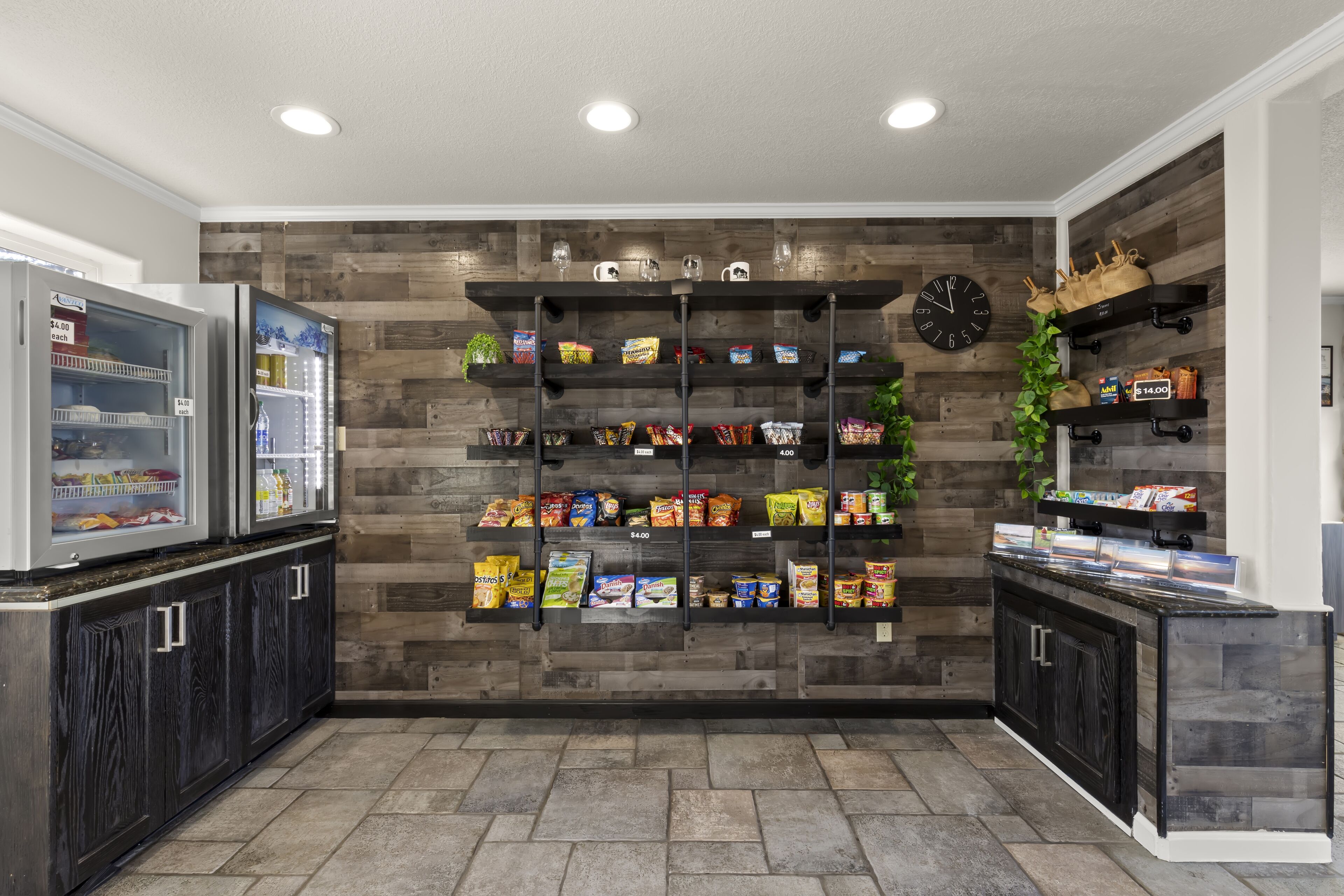 A kitchen with a wooden wall, a clock, and shelves with snacks and beverages.