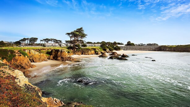 A coastal landscape with a sandy beach, rocky shoreline, and a pier extending into the water.
