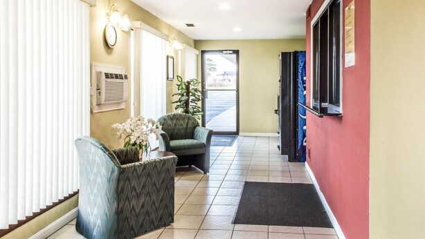 A reception area with a clock, air conditioning unit, two armchairs, a potted plant, and a door leading outside. - Rodeway Inn Shippensburg I-81 (Shippensburg)