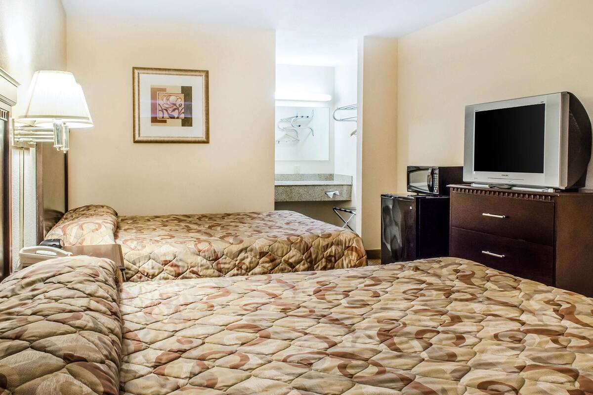 Une chambre d’hôtel avec deux lits, une télévision, un bureau avec une lampe, un tableau encadré au mur et une salle de bain visible à travers une porte ouverte.