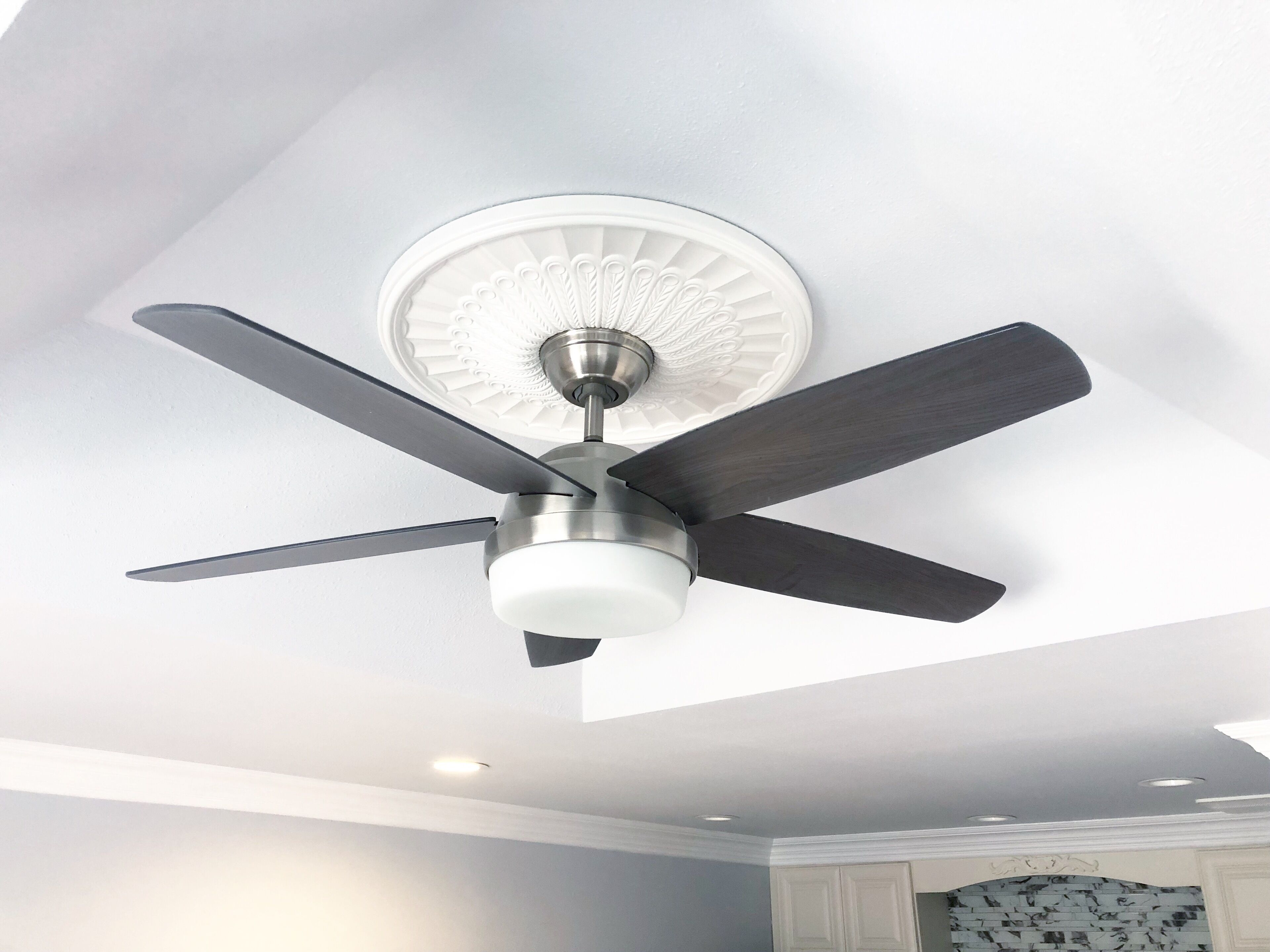 Um ventilador de teto com quatro lâminas e uma luminária central em um quarto com paredes brancas e um design decorativo no teto.