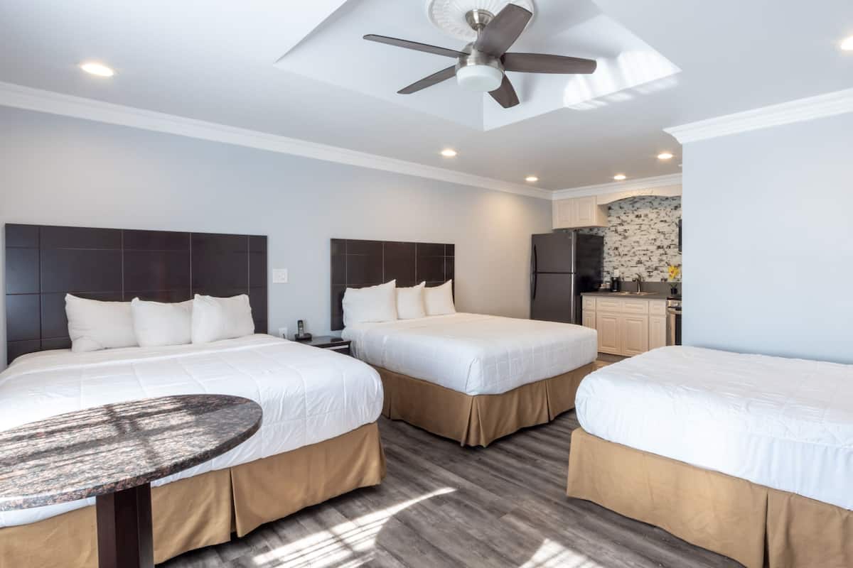 Ein Hotelzimmer mit drei Betten, einem Deckenventilator, einem kleinen Tisch und einer Küchenzeile im Hintergrund.