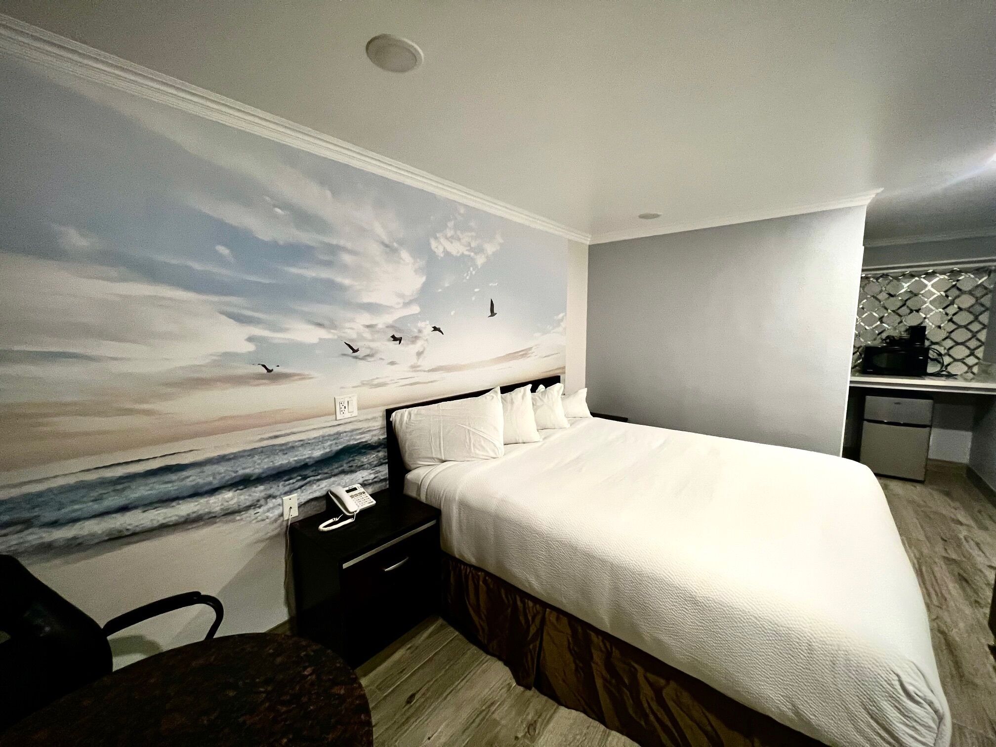 A hotel room with a large bed, a nightstand with a phone, a small desk with a microwave, and a wall mural of a beach scene.