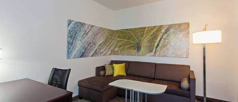 A modern hotel room with a brown sectional sofa, a small white table, a dark wooden desk, a floor lamp, and a large abstract painting on the wall.