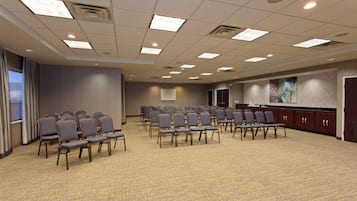 Un salón de conferencias con filas de sillas, una pantalla de proyector y una zona de kitchenette.