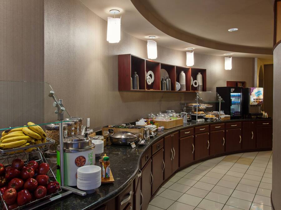 Zona de desayuno de hotel con barra de buffet, que incluye frutas, bebidas y diversos alimentos.