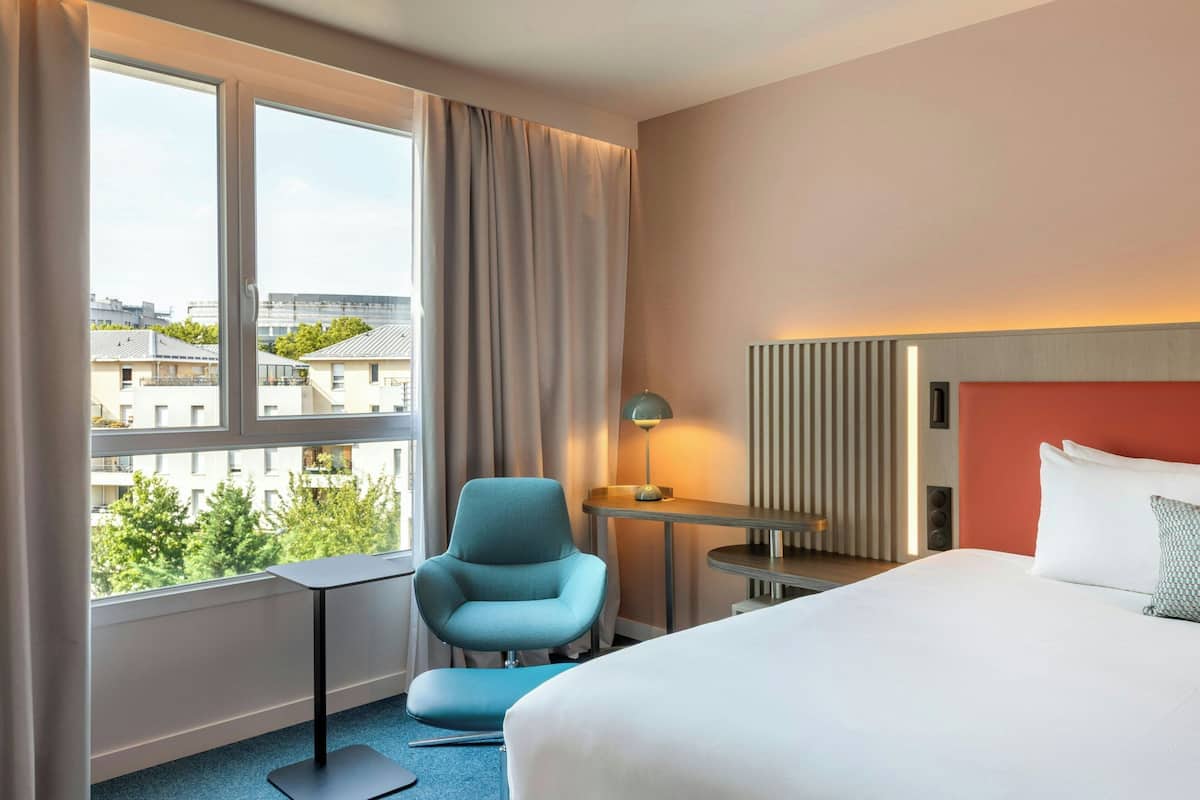 A hotel room with a bed, a desk, a chair, a window with curtains, and a view of buildings.