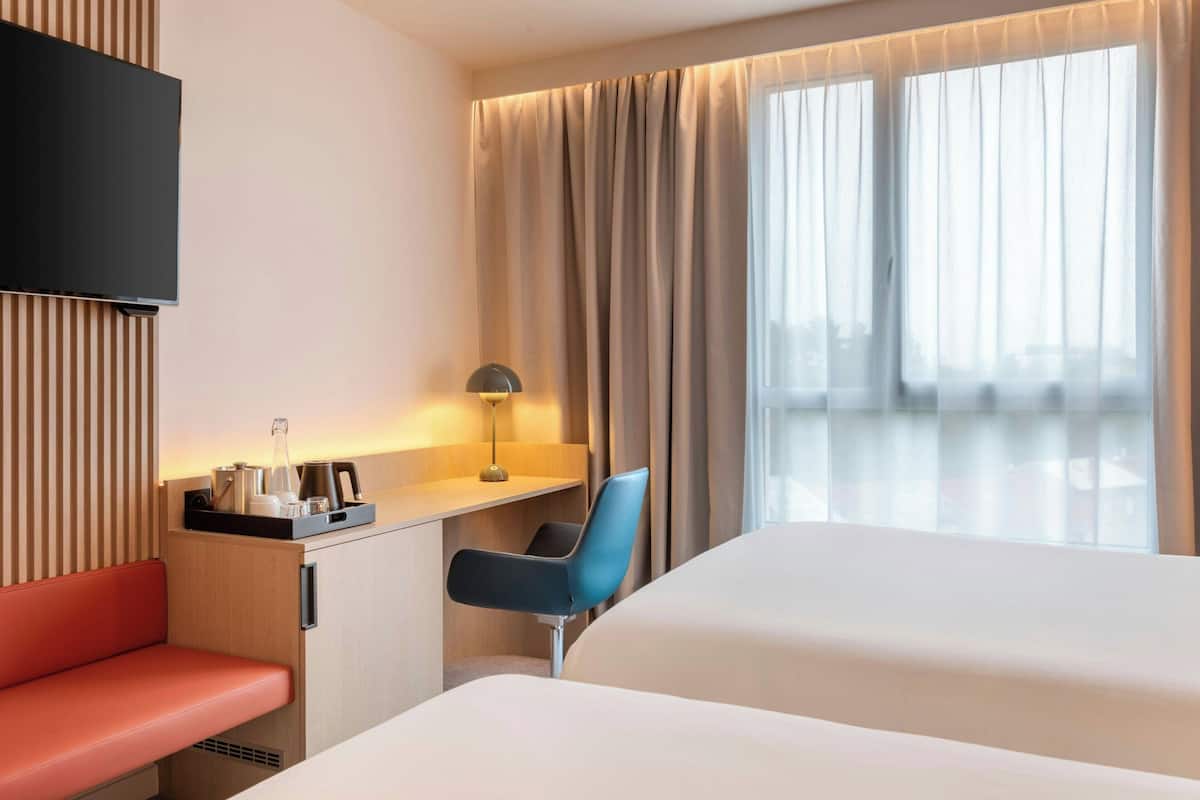 A hotel room with a bed, a desk with a chair, a TV mounted on the wall, and a window with curtains.