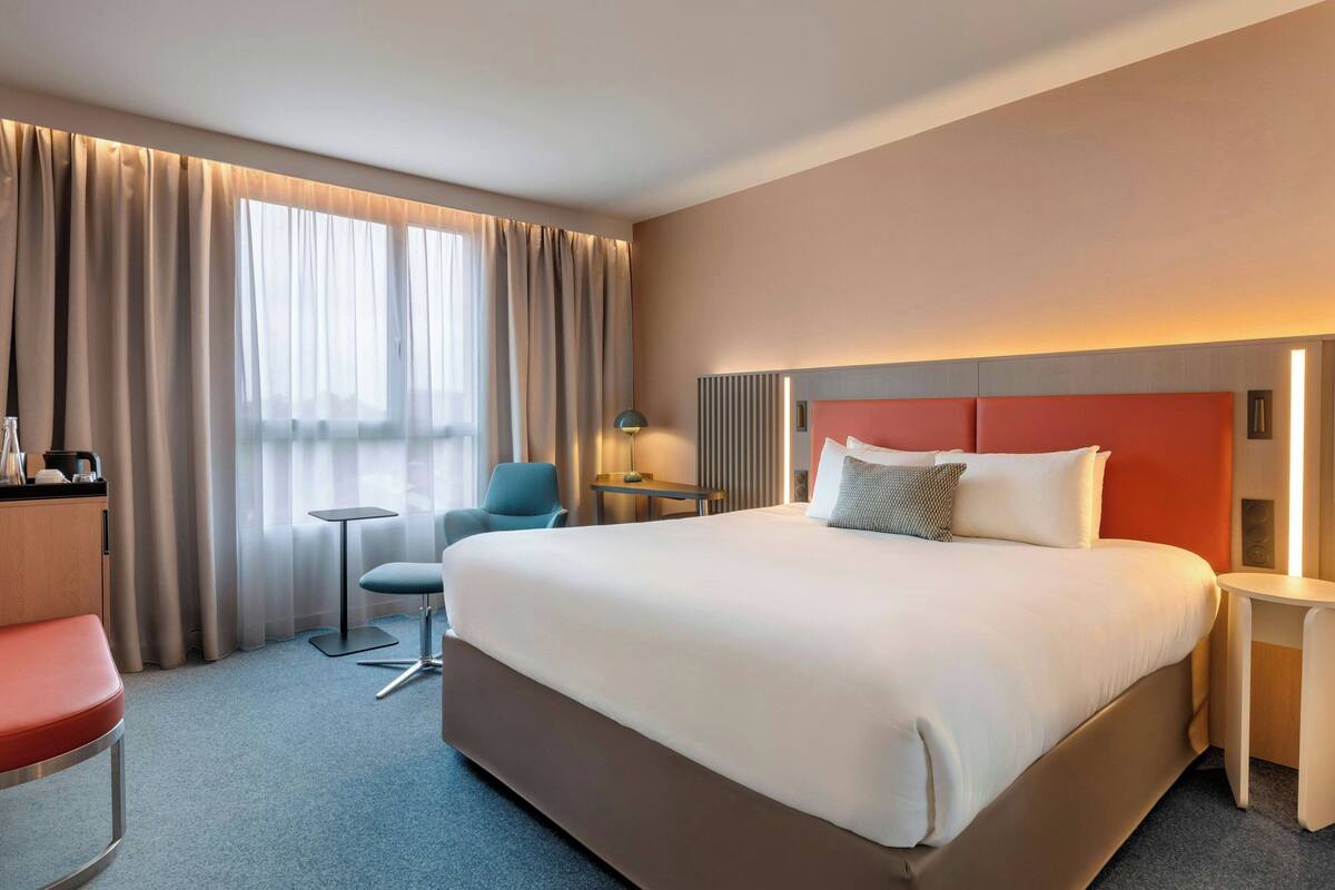 A hotel room with a large bed, a desk, a chair, and a view of the city through the window.