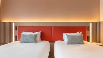 Habitación de hotel con dos camas, cada una con ropa de cama blanca y almohadas estampadas. Los cabeceros son rojos, y hay una tira de luz sobre las camas.
