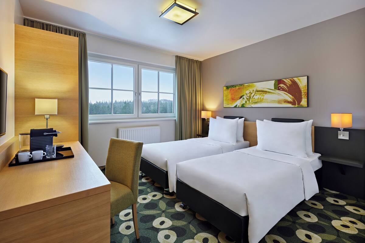 A hotel room with two beds, a desk with a lamp, a chair, and a window with a view of trees.