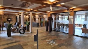 A spacious lobby with a stone wall, wooden beams, and a seating area with chairs and potted plants. - Nürburgring Motorsport Hotel (Nuerburg)