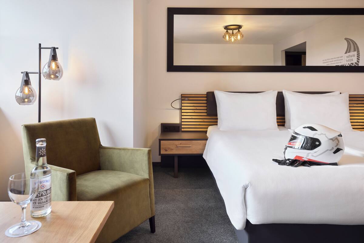 A hotel room with a bed, a chair, a nightstand, a lamp, and a bottle of alcohol on a table.