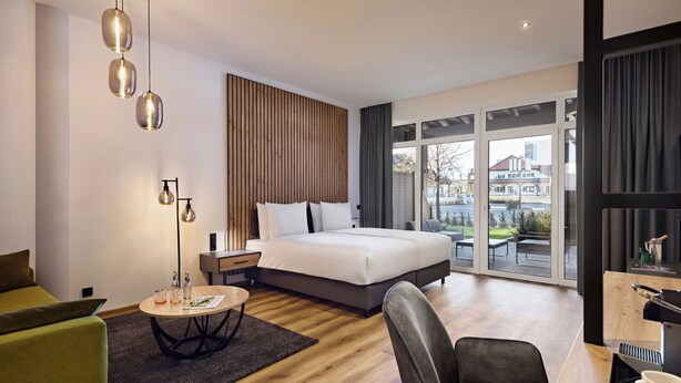 A modern hotel room with a large bed, a desk with a coffee machine, a green sofa, and a view of the city through large windows. - Nürburgring Motorsport Hotel (Nuerburg)