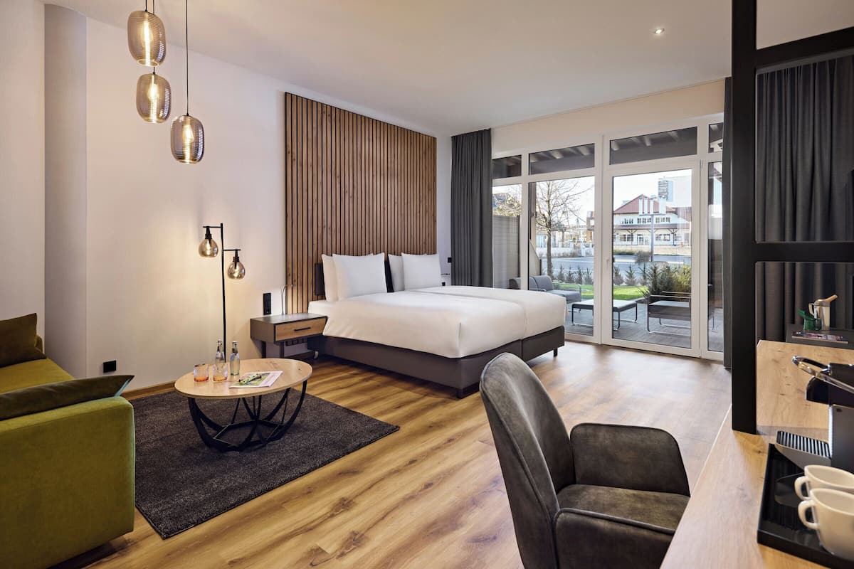 A modern hotel room with a large bed, a desk with a coffee machine, a green sofa, and a view of the city through large windows.