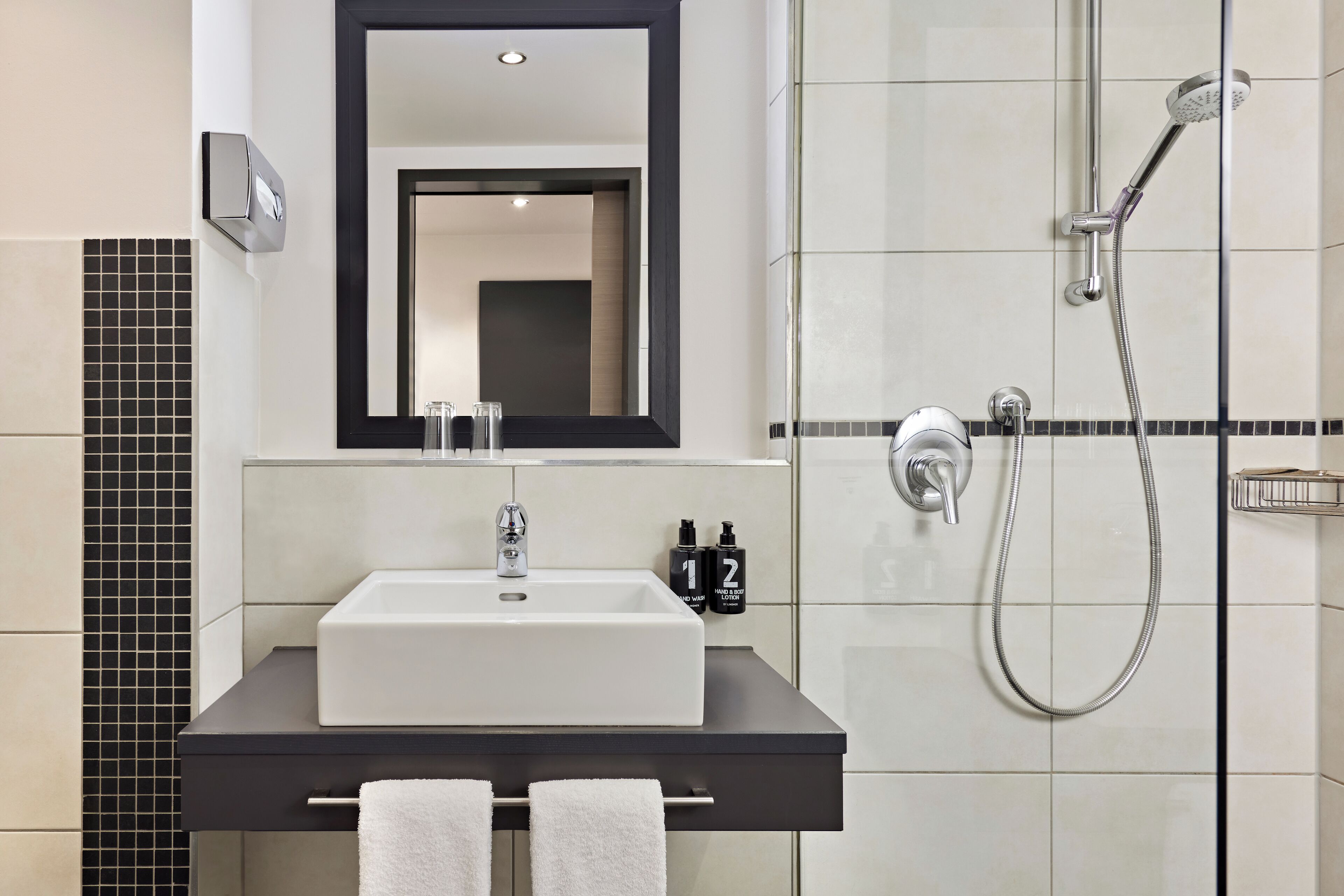 Ein modernes Badezimmer mit einem weißen Waschbecken, einer schwarzen Arbeitsplatte und einem Spiegel darüber. Auf der Arbeitsplatte stehen zwei Flaschen Hand- und Körperlotion.