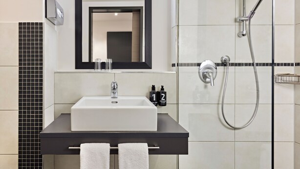 A modern bathroom with a white sink, black countertop, and a large mirror. There is a showerhead and a towel rack visible. - Nürburgring Motorsport Hotel (Nuerburg)