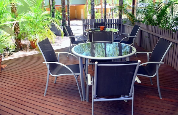 A round glass table with six chairs, surrounded by a wooden deck and potted plants.